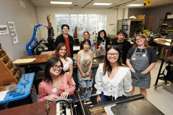 A group of smiling students pose together in a printmaking studio surrounded by printing presses, type trays, and tools. The environment is warm and collaborative, highlighting a hands-on, creative learning space.