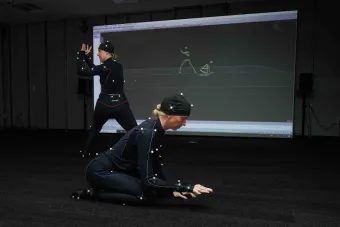 Two performers in motion-capture suits demonstrate choreography in a dark studio, with their movements digitally rendered in real time on a large screen behind them. The space highlights immersive technology and interdisciplinary collaboration.