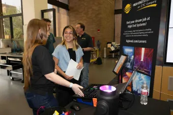 A student smiles while speaking with a representative from High End Systems at a career event booth. Behind them is a large promotional display advertising careers in lighting technology for the entertainment industry.