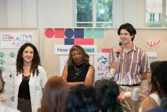 Three faculty members stand at the front of a classroom-style event, engaging with a seated audience. One holds a microphone while speaking, and illustrated signage with phrases like “How to Interact” and “Collect and Scan at kiosk” appears in the background.