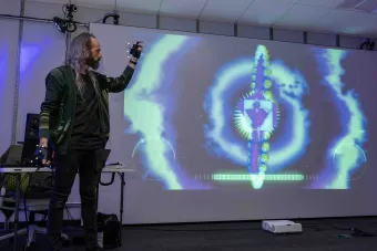 A faculty member wearing motion-tracking gloves demonstrates immersive technology in front of a large projection screen. The screen displays an abstract, colorful digital interface resembling a cosmic visualization. Cables, equipment, and a projector are visible in the space.