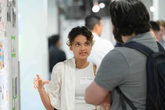A student wearing a name tag that reads “Maya Lamari” presents her design work to an attendee at an exhibition. She gestures with her hands while speaking, standing beside a wall covered in printed visual materials. The background is softly blurred, showing other people engaged in conversation.