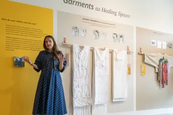 A woman stands in front of a wall display titled "Garments as Healing Spaces," presenting a collection of sensory and symbolic garments. She gestures with open hands while standing beside white fabric prototypes hung on dowels and large descriptive text panels explaining the emotional and symbolic functions of clothing.