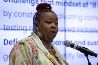 A speaker in a floral blazer addresses an audience from a microphone. Behind her, bold projected text highlights design goals such as inclusivity, testing, and transparency. She wears statement earrings and a layered necklace.
