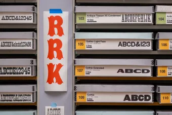 Close-up of labeled type drawers in a print lab. A bold red poster in the center reads “RRK” in a decorative font. Drawers surrounding the poster are labeled with typeface names like “Gothic Light Face,” “Antique Tuscan XX Condensed,” and “Gothic Extended,” each showing example characters.