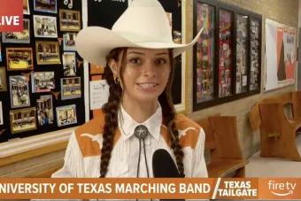 Student in western attire with braided hair and a white cowboy hat being interviewed live on TV about the University of Texas Marching Band. A bulletin board with Longhorn-themed photos and memorabilia appears behind her.