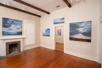 Interior view of Verdant Gallery showing landscape and cloudscape paintings by AET faculty member Karen Maness. The room features cream-colored walls, wood floors, and exposed ceiling beams with track lighting.