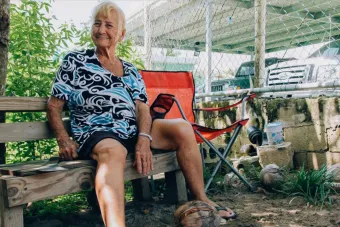 An older woman with short white hair sits on a wooden bench outdoors, smiling warmly. She wears a black and white patterned shirt and black shorts, resting her hand on a coconut at her feet. A red folding chair, cinder blocks, and parked cars are visible behind a chain-link fence in the background.