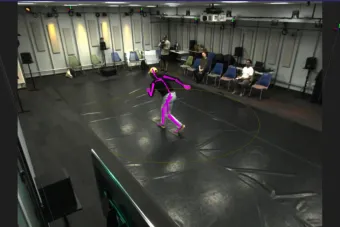 A person performs motion capture movements inside a circular boundary in a high-tech lab space. A bright pink digital skeleton overlay tracks their posture in real-time. Seated observers watch from the edge of the room, while cameras and sensors are mounted along the walls and ceiling.