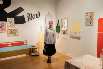 A smiling woman stands in the center of a gallery-style installation featuring playful, illustrated wall art and hand-crafted graphic design objects. Framed prints, textiles, and printed materials surround her, including a series titled “No Phone Zone.” The immersive space blends printmaking, design, and everyday objects into a cohesive visual narrative.