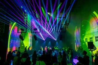 A large crowd gathers in a dark performance space illuminated by vivid green, blue, and magenta laser beams that stretch across the room. DJs perform at tables along the wall while animated projections wash the walls in circular patterns, creating an immersive audiovisual experience that blends music, light, and digital art.