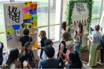 Students gather around exhibition boards displaying design projects titled “Unfolded” and “Back to the Roots.” Colorful printed spreads, books, and posters are mounted on the walls, while attendees converse, point to details, and scan a QR code. The bright, window-lined space emphasizes collaboration, research, and presentation in a design showcase setting.