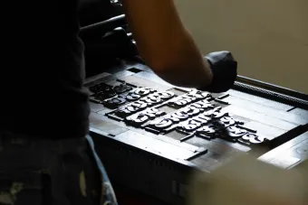 A person wearing a protective glove arranges wood type blocks on the bed of a letterpress. The dark metal and carved letterforms are set in rows, capturing the hands-on process of composing type for print in a design lab setting.