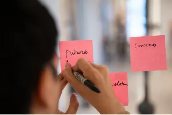 A person writes the word "Future" on a pink sticky note affixed to a glass surface, with additional sticky notes nearby during a design thinking activity.