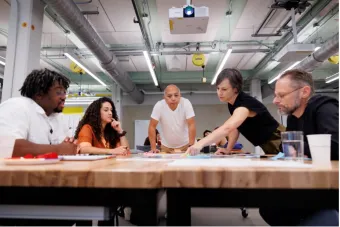 Five people gathered around a worktable in a studio space, collaborating over printed materials and colorful sticky notes.