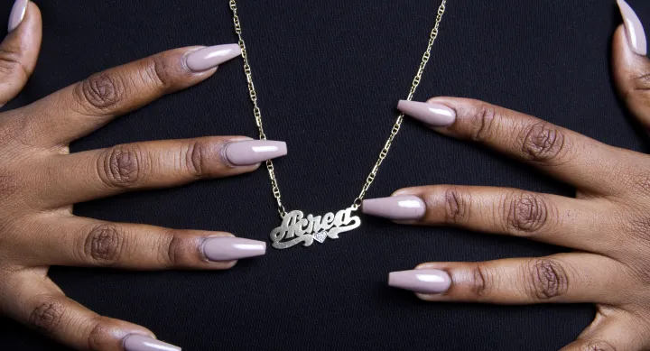 A close-up photograph of a person’s hands with long, glossy, nude-colored nails gently holding a gold nameplate necklace that reads 'Acead' in script font. The necklace has a small heart detail and hangs against a black fabric background.
