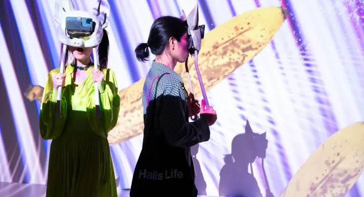 Two people participate in an immersive experience wearing virtual reality headsets mounted on animal-shaped masks, holding poles for support. Vivid digital projections of bananas and streaks of light animate the space around them, casting shadows on the wall.