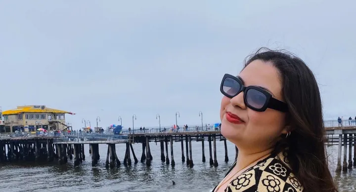 Xandria Hernandez wearing a patterned cream and black cardigan standing by the ocean with a wooden pier in the background under a cloudy sky.