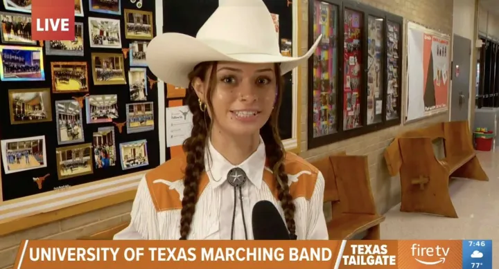 Student in western attire with braided hair and a white cowboy hat being interviewed live on TV about the University of Texas Marching Band. A bulletin board with Longhorn-themed photos and memorabilia appears behind her.