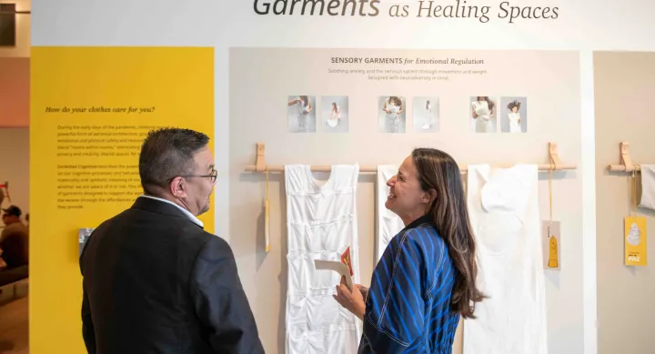 A woman in a blue striped dress speaks with a man in front of an exhibition display titled "Garments as Healing Spaces." The exhibit features white sensory garments designed for emotional regulation and panels explaining the concept of clothing as personal architecture for mental and emotional care.
