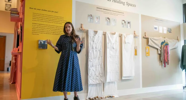 A woman stands in front of an exhibition titled "Garments as Healing Spaces," gesturing as she speaks. Behind her, white sensory garments for emotional regulation are displayed on a wall alongside descriptive text panels and photos illustrating their use.