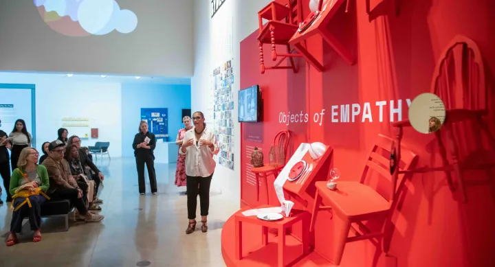 A speaker presents to a seated audience in front of a bright red installation titled “Objects of Empathy,” featuring chairs, tables, and household items mounted to the wall. The exhibit is part of a design showcase exploring empathy through everyday objects.