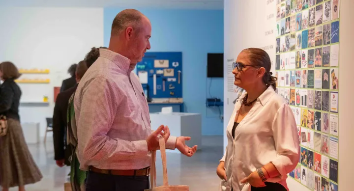 Two attendees talk during an MFA info session, standing in front of a wall filled with colorful graphic design tiles. The interaction takes place in a well-lit gallery space with other guests exploring the exhibition in the background.