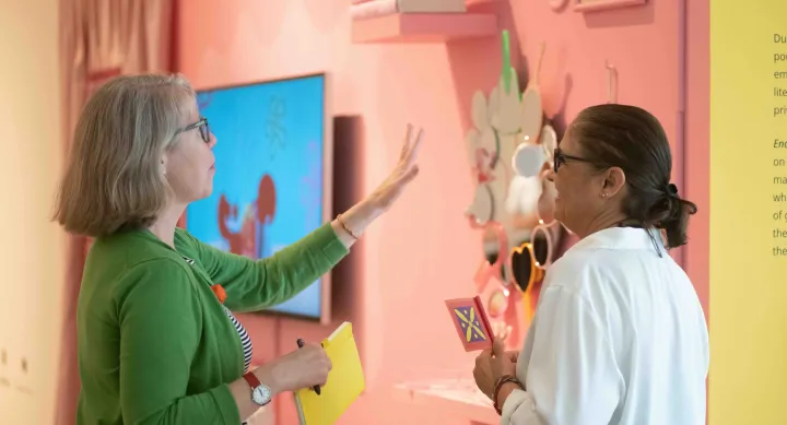 Two attendees chat in front of a pink wall display featuring colorful mirrors and floating books. One gestures with her hand while holding a notebook, and the other smiles while holding a brightly designed card.