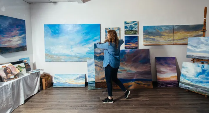 Artist arranging large landscape paintings in a studio gallery space, surrounded by vivid skies and cloudscapes displayed on easels and walls.