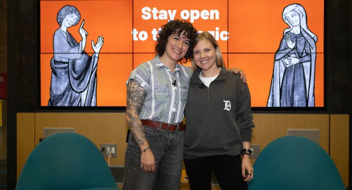 Two smiling speakers stand arm-in-arm in front of a large digital display with the words “Stay open to the magic” and medieval-style illustrations of gesturing figures.