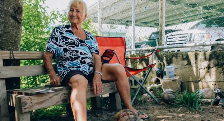 An older woman with short white hair sits on a wooden bench outdoors, smiling warmly. She wears a black and white patterned shirt and black shorts, resting her hand on a coconut at her feet. A red folding chair, cinder blocks, and parked cars are visible behind a chain-link fence in the background.