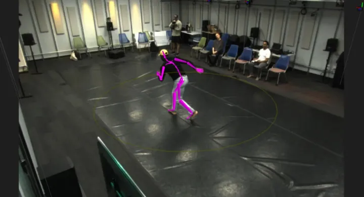 A person performs motion capture movements inside a circular boundary in a high-tech lab space. A bright pink digital skeleton overlay tracks their posture in real-time. Seated observers watch from the edge of the room, while cameras and sensors are mounted along the walls and ceiling.