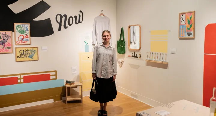 A smiling woman stands in the center of a gallery-style installation featuring playful, illustrated wall art and hand-crafted graphic design objects. Framed prints, textiles, and printed materials surround her, including a series titled “No Phone Zone.” The immersive space blends printmaking, design, and everyday objects into a cohesive visual narrative.