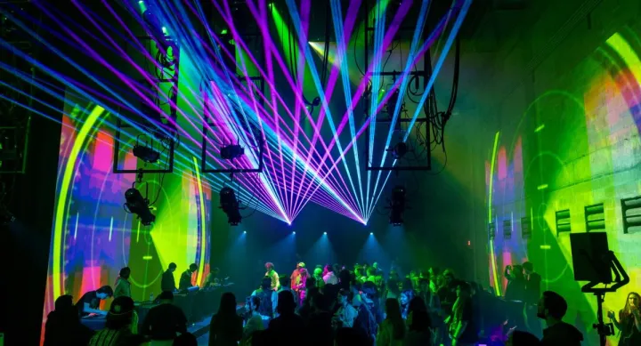 A large crowd gathers in a dark performance space illuminated by vivid green, blue, and magenta laser beams that stretch across the room. DJs perform at tables along the wall while animated projections wash the walls in circular patterns, creating an immersive audiovisual experience that blends music, light, and digital art.