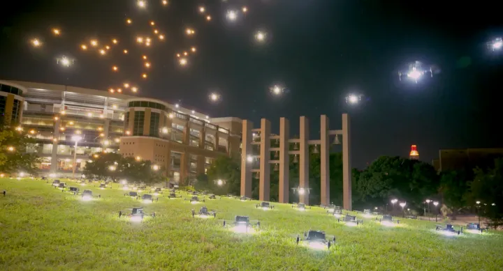 Dozens of drones line a grassy hillside on the University of Texas at Austin campus at night, with more drones ascending into the sky above university buildings and the Tower glowing in the distance.
