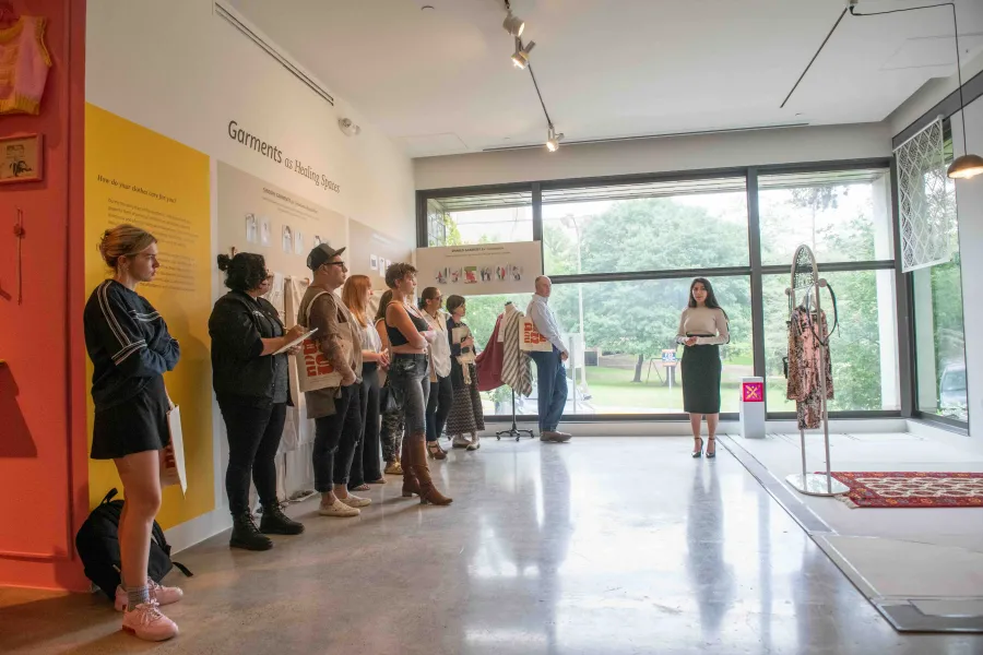 A group of people stand in a gallery space listening to a woman speak near large windows. The exhibition, titled "Garments as Healing Spaces," features text panels, sensory garments, and clothing installations focused on emotional regulation and care.