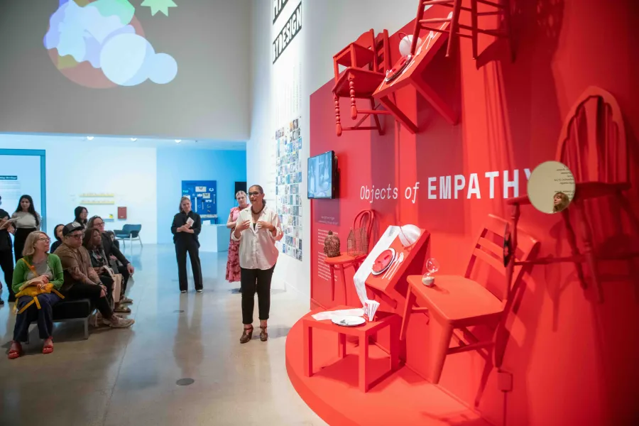 A speaker presents to a seated audience in front of a bright red installation titled “Objects of Empathy,” featuring chairs, tables, and household items mounted to the wall. The exhibit is part of a design showcase exploring empathy through everyday objects.