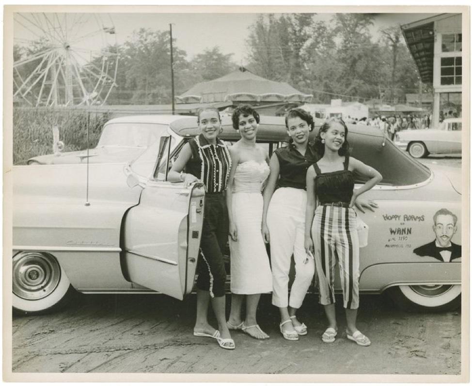 1960s Black and white photo of four Black women standing outside of a classic car at a carnival