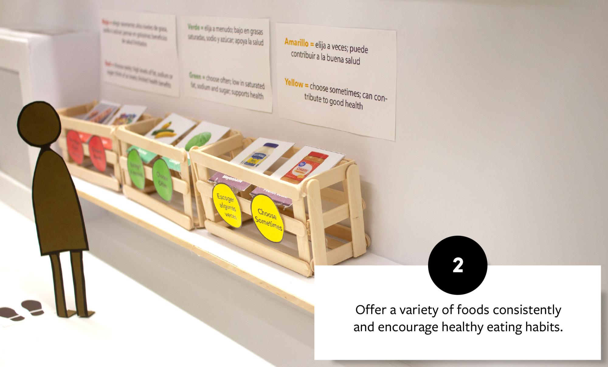 Cardboard figure stands in front of a display of labeled food bins, promoting healthy eating choices, with signs in Spanish and English encouraging balanced dietary habits.