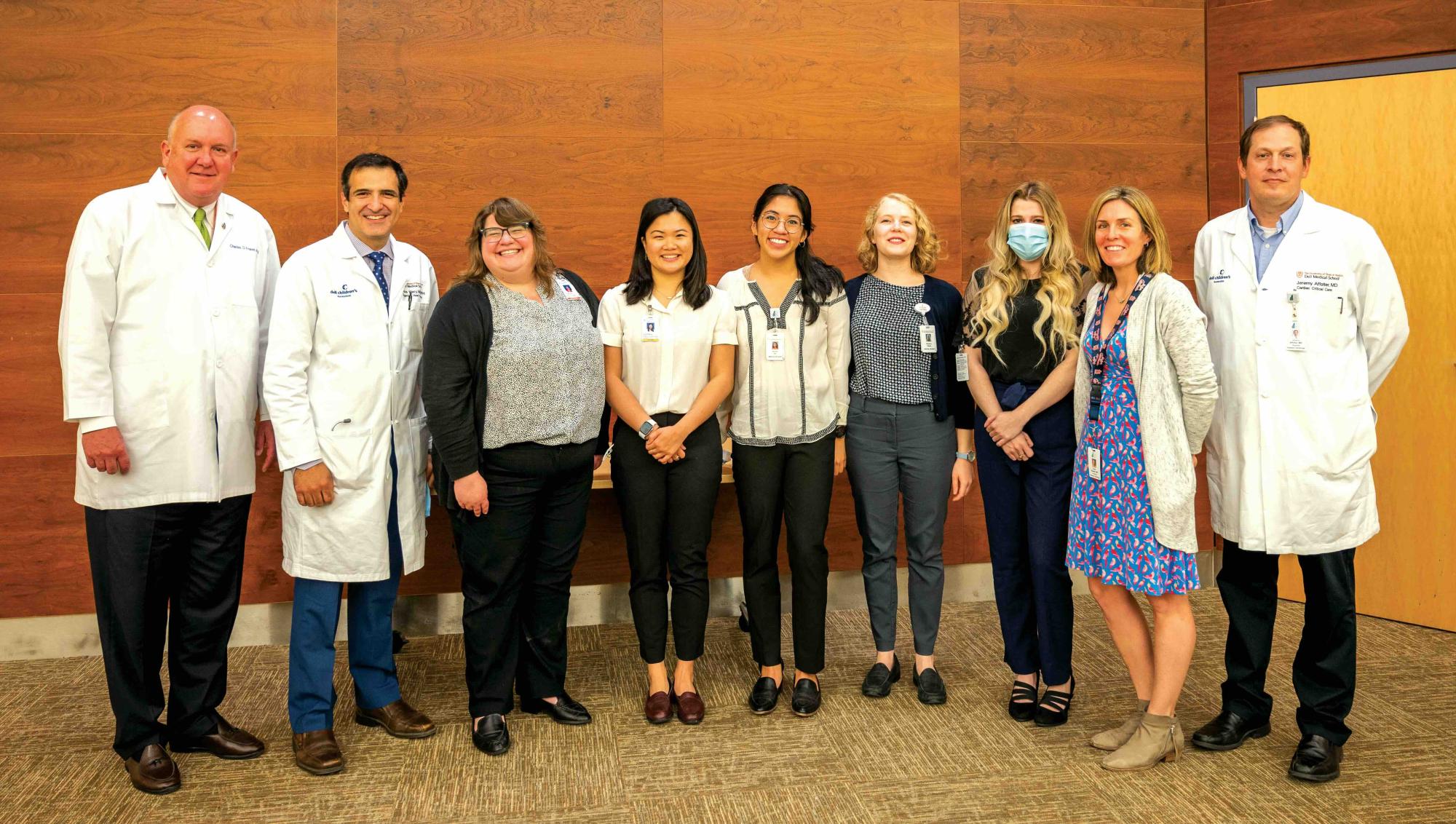 A group of ten healthcare professionals stands in a line smiling for a group photo in front of a wooden wall. The team includes individuals in white lab coats, business attire, and clinic uniforms. One person wears a medical mask, and several are wearing ID badges. The setting appears to be a hospital or medical facility, with carpeted floors and a professional yet welcoming atmosphere.