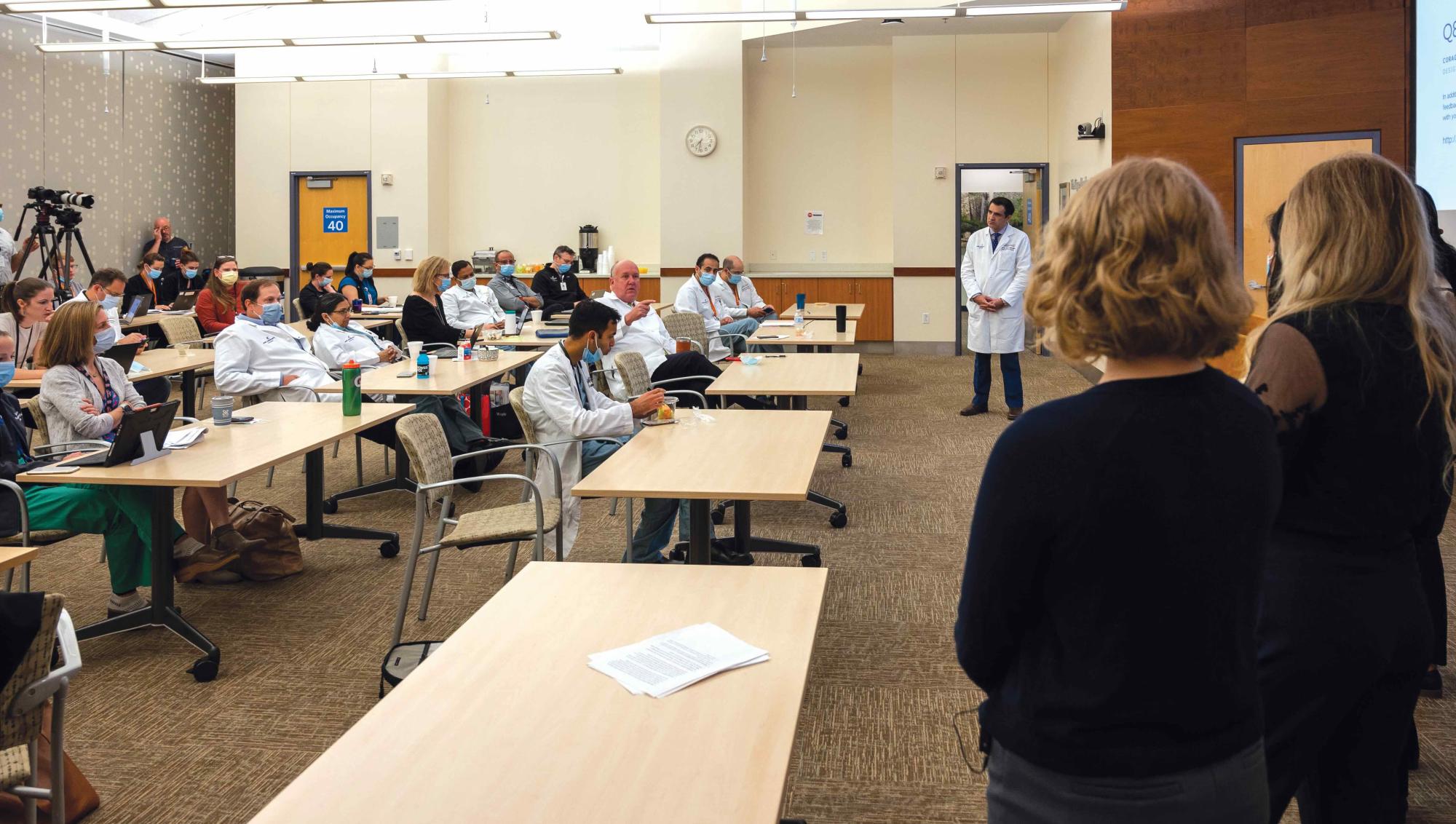 A packed conference room filled with medical professionals seated at long tables, attentively listening to a presentation. Many attendees wear white lab coats and surgical masks, indicating a healthcare setting. Two presenters stand at the front facing the audience, while a third person in a lab coat stands near the screen. The space is well-lit, with cameras positioned in the back capturing the event.