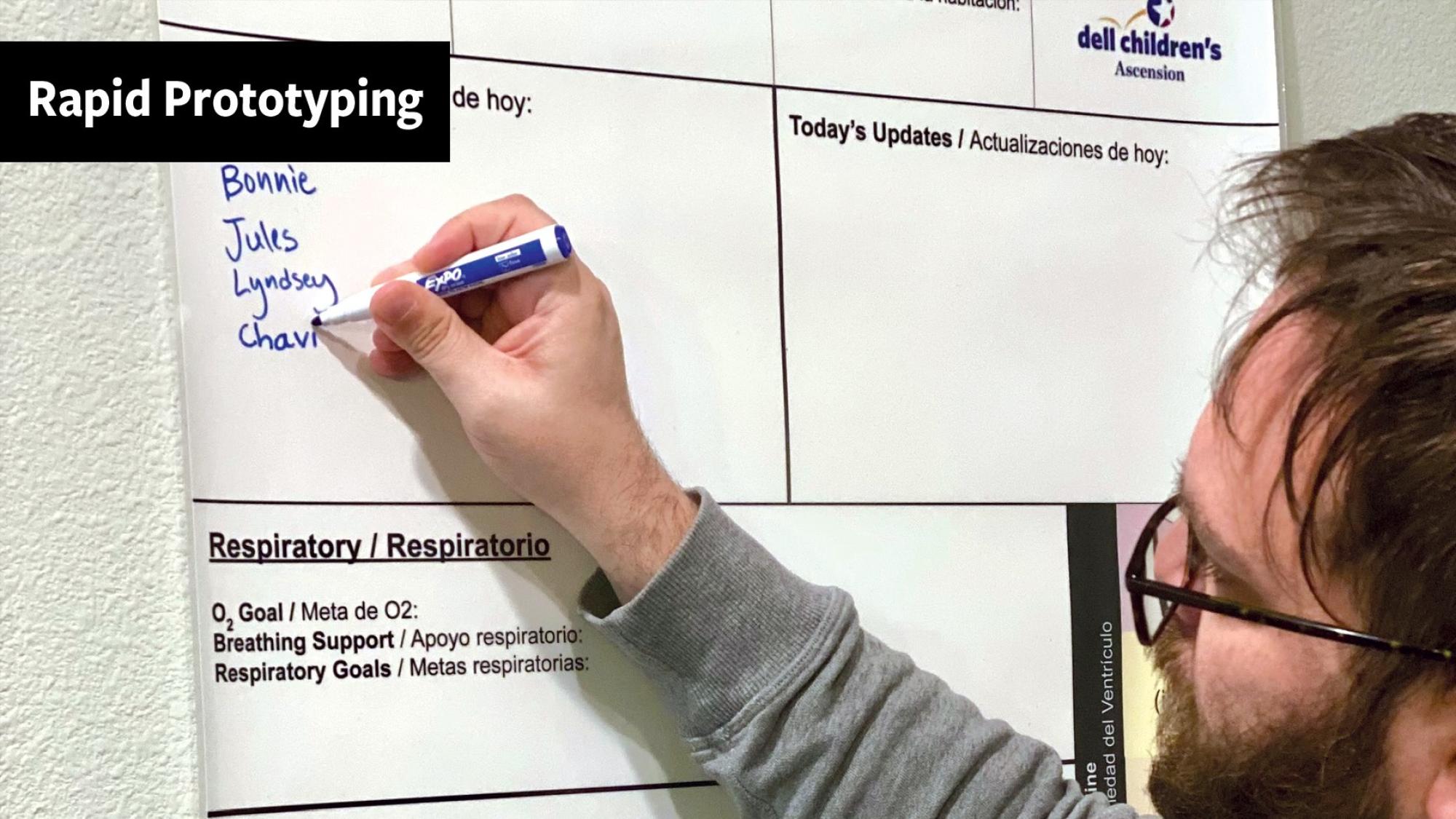 A person with glasses writes staff names on a dry-erase prototype care board mounted on a wall. The board, branded with the Dell Children’s Ascension logo, includes bilingual sections such as “Today’s Updates / Actualizaciones de hoy” and “Respiratory / Respiratorio.” A black banner in the upper left corner reads “Rapid Prototyping.”