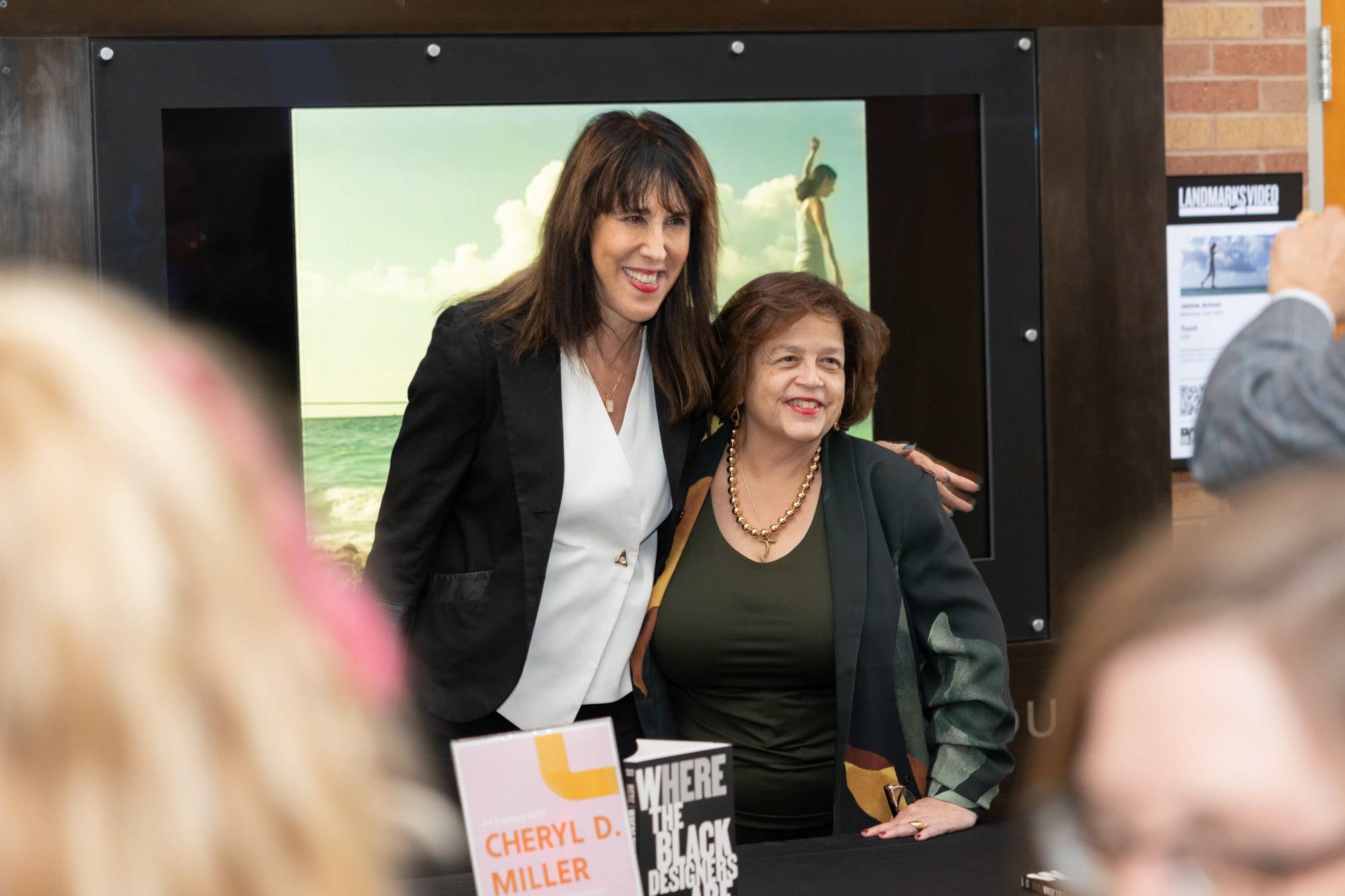Two individuals pose for a photo during a book signing event. They are standing in front of a framed image of a beach scene, with copies of Cheryl D. Miller’s book Where Are the Black Designers? visible on the table in the foreground.