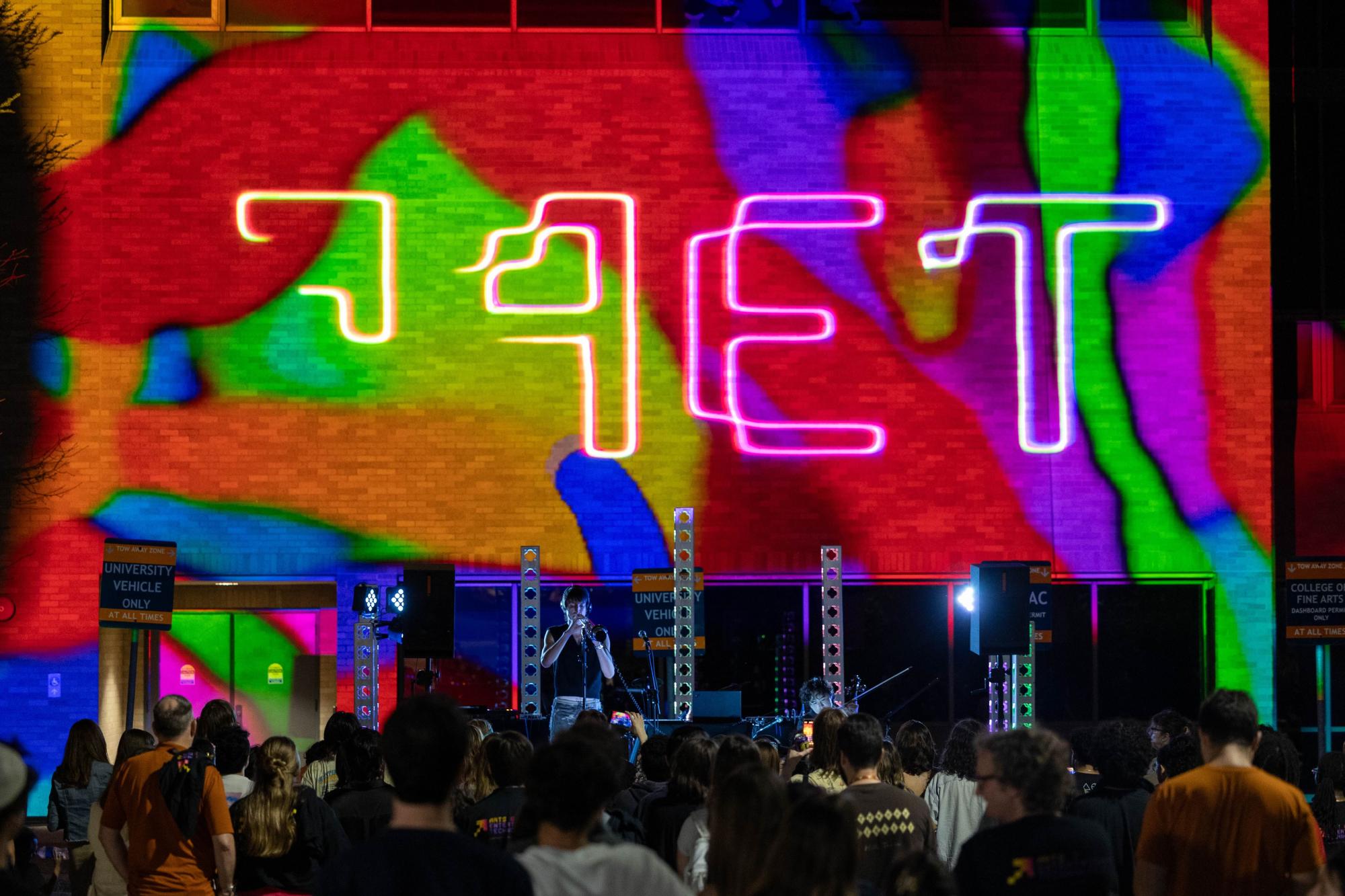 A large crowd gathers outside at night, facing a colorful projection on a brick building that reads "AET" in glowing neon text. The vivid background features swirling, abstract rainbow patterns. A performer stands center stage under the projection, illuminated by concert lights, with musical equipment and speakers set up around them.