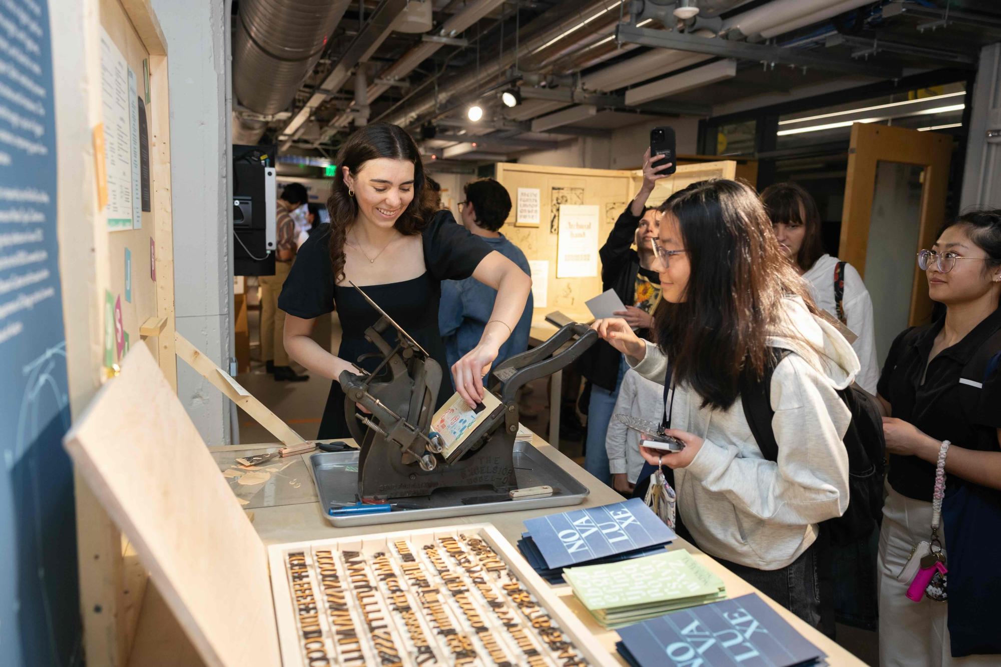 A student demonstrates a letterpress printing process at an event table, guiding a guest who is pressing a design onto a card. Attendees observe and wait in line, with printed materials and type blocks displayed in the foreground.