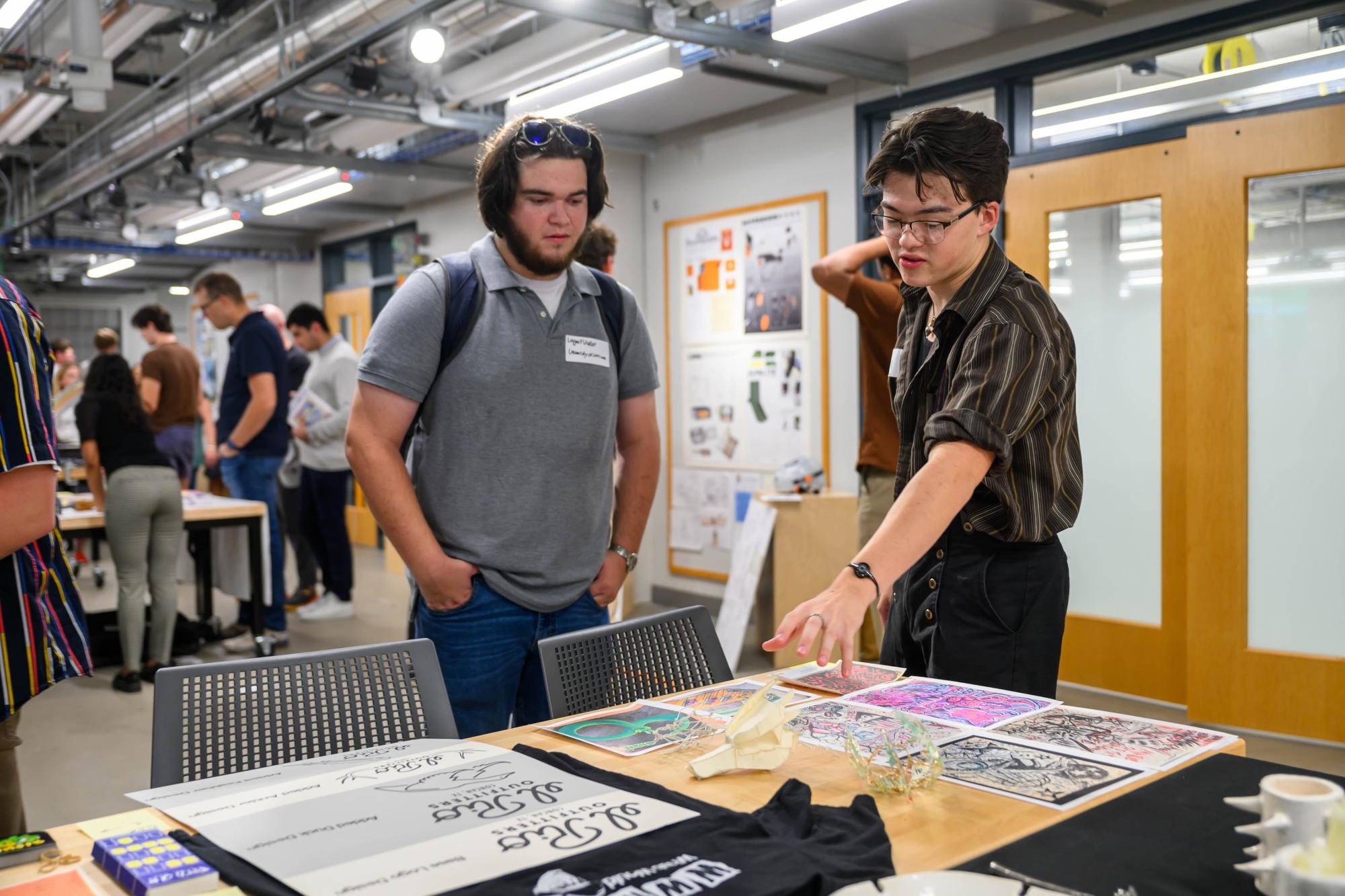 A student explains their printmaking and illustration work to a visitor at a showcase event. Prints, t-shirts, and other creative pieces are displayed on a table in front of them. Other attendees interact in the background of the studio space.