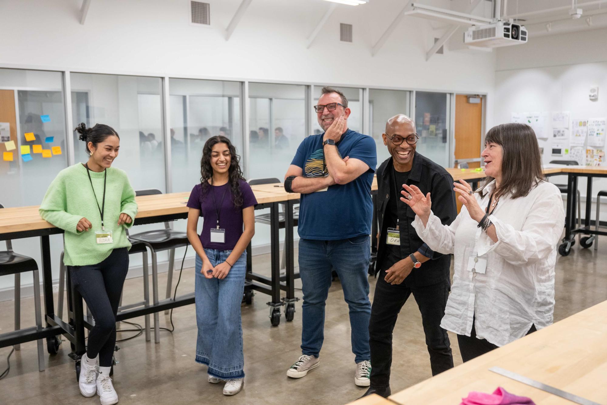 A group of five people—two students and three instructors—stand together in a design studio classroom, smiling and laughing during a collaborative activity. Large wooden tables and glass-walled meeting rooms are visible in the background.