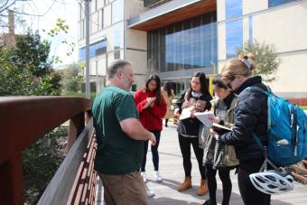 Design students stand on a bridge outside and take notes as they study the creek below them and their professor lectures