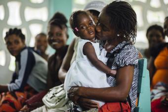 a Haitian mother holds her young daughter, who wears a white dress and is looking directly into the camera