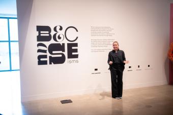 Graduate Program faculty Kelcey Gray standing in front of the Becauseisms logo and exhibition description